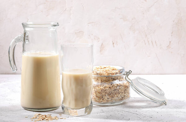 Transparent decanter and glass of oat milk and jar with oat flakes on white background.