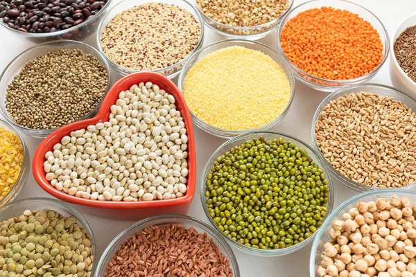 Close-up glass bowls with cereals, beans and seeds with red heart-shaped bowl in the middle on white background.