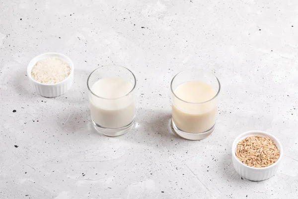 Glasses with rice and oat milk, bowls with rice seeds and oat flakes on concrete background.
