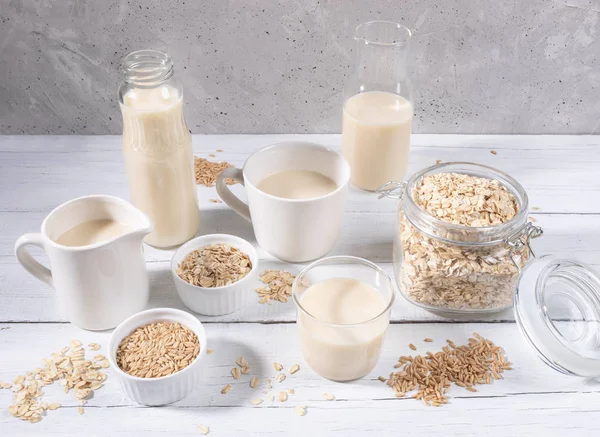 Set of different containers with oat milk, oat seeds and flakes on white wooden table on concrete background.