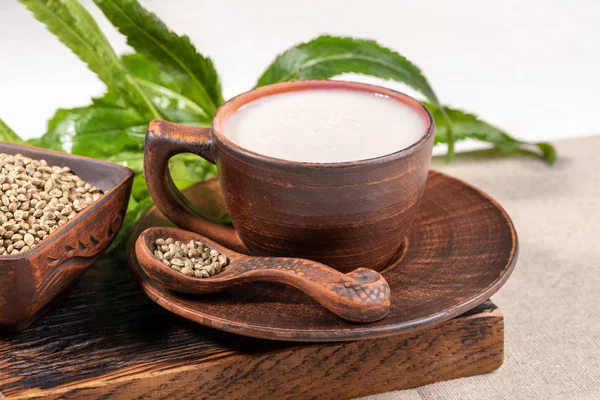 Hemp milk in ceramic cup and grains in brown ceramic bowl and spoon and cannabis leaves.