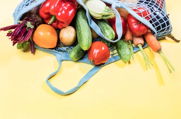 Blue mesh shopping bag full of fresh farm vegetables is lying on top of yellow background with copy space on bottom. — Stock Photo, Image