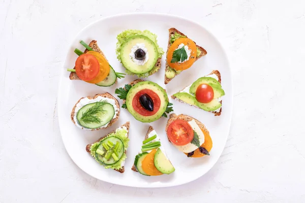 Set of vegetarian sandwiches with tomatoes, avocado, cucumber, greenness, cheese and sun-dried olives on white plate in center of white textured background. Top view, flat lay, copy space.