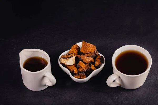 Two white ceramic cups of healing infusion of birch mushroom chaga with chaga pieces in bowl on black backdrop. Chaga mushroom coffee or tea. This drink rich by antioxidants for immunity. Copy space.