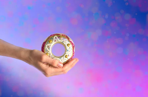 Mulher mão segurando delicadamente brinquedo de Natal - decorativo donut de vidro colorido no fundo multicolorido rosa-roxo . — Fotografia de Stock
