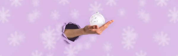 白いクリスマスボールを持っている女性の手と広いバナーを持っています引き裂かれた穴を通ってピンクの背景の真ん中に雪の結晶. — ストック写真