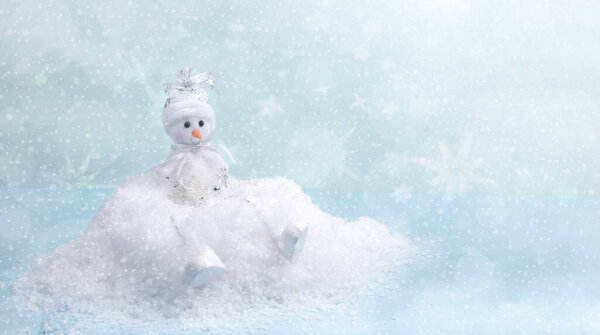 Xmas greeting card. Cute small snowman sitting on white snowdrift on light blue backdrop with snow, stars and bokeh.