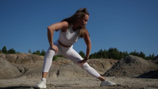 Chica rubia en traje blanco hace estiramiento piernas rodeada de arenas — Vídeos de Stock