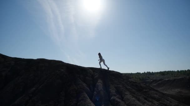 La silueta de una chica que entrena en una colina contra un cielo azul — Vídeos de Stock