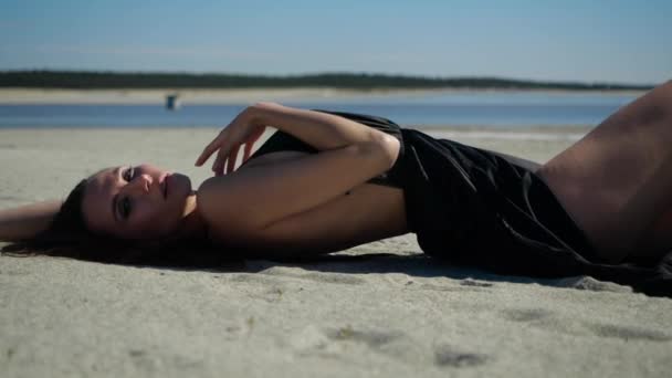 Eine junge Frau im schwarzen Kleid liegt im Sand und macht verführerische Bewegungen — Stockvideo