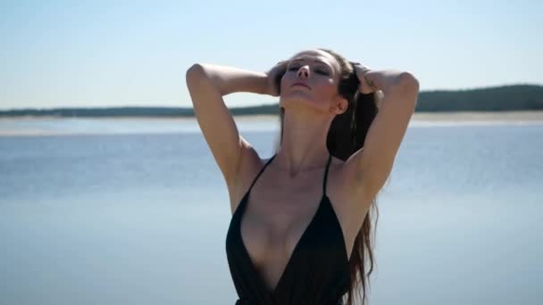 Струнка дівчина в чорному літньому платті вирівнює волосся на пляжі — стокове відео