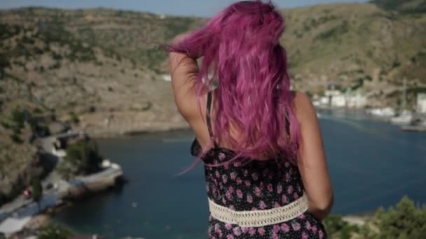 Девушка с взъерошенными розовыми волосами в платье с цветочным отпечатком смотрит на воду — стоковое видео