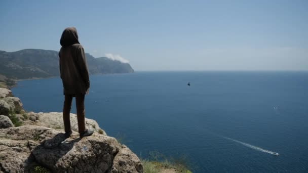 Un hombre en una capucha en el borde del acantilado frente a él tiene una vista del mar — Vídeos de Stock