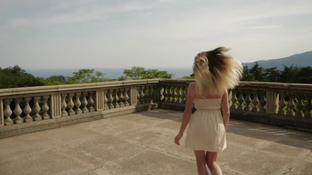 Chica en vestido blanco en una amplia terraza antigua mira a los árboles — Vídeo de stock