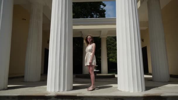 Una mujer con un vestido blanco posa de pie entre dos altas columnas blancas — Vídeos de Stock