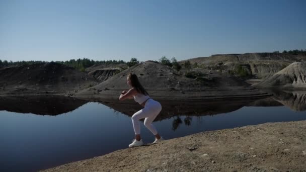 Mujer en un chándal blanco realiza sentadillas con saltos en la orilla del lago — Vídeos de Stock