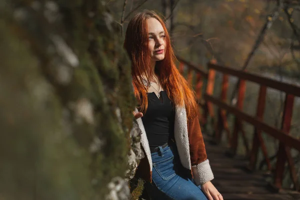 Een meisje met rood haar op de brug, leunend tegen een rots, starend naar de zon — Stockfoto