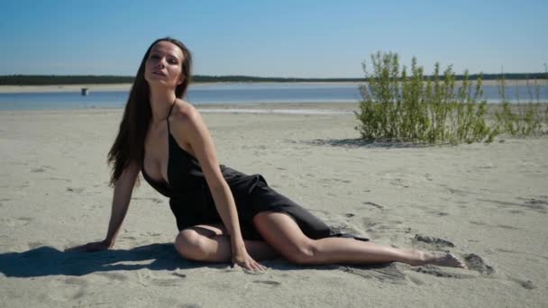 Affascinante ragazza in abito nero leggermente coprendo corpo nudo si siede sulla spiaggia — Video Stock