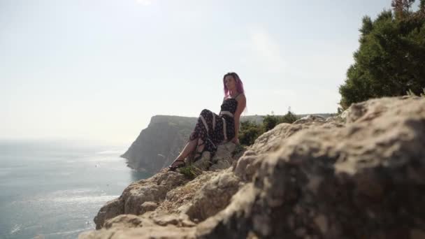 Λεπτή κοπέλα σε ένα μακρύ φόρεμα με ροζ μαλλιά κάθεται στην άκρη του γκρεμού — Αρχείο Βίντεο