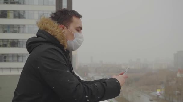 Um cara com uma máscara médica e um sobretudo está se apoiando no corrimão do terraço — Vídeo de Stock