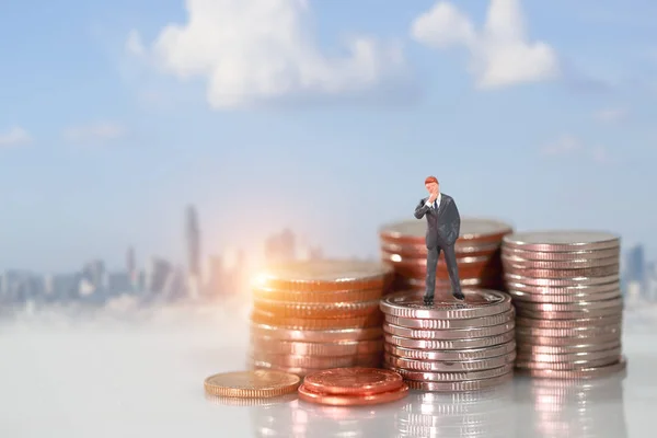 Personas en miniatura: hombre de negocios de pie en el podio de apilamiento de monedas con fondo borroso de la ciudad (esta imagen para el concepto de competencia financiera y empresarial — Foto de Stock