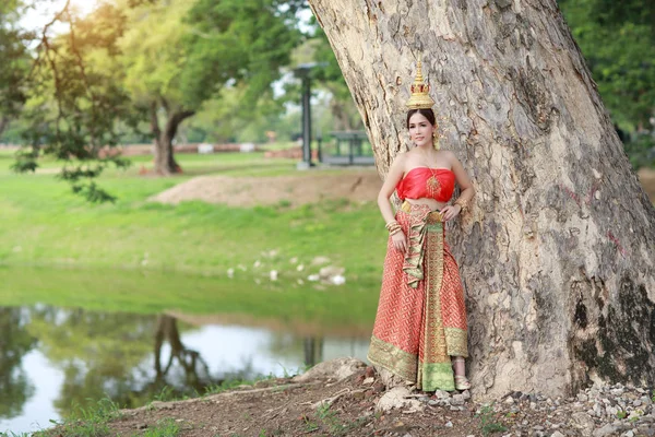 Young asian fashion girl wearing Thai traditional costume with beauty face standing beside big green trees in nature outdoors (using for background) — Stock Photo, Image