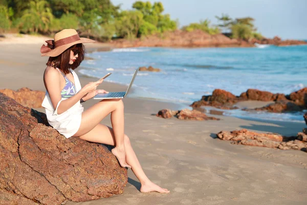 美丽可爱的女商人使用笔记本电脑和手机与太阳眼镜和帽子在海滩上度假 — 图库照片