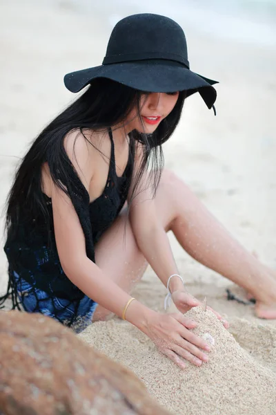 美丽的女人坐在沙滩上玩沙与黑帽子 — 图库照片