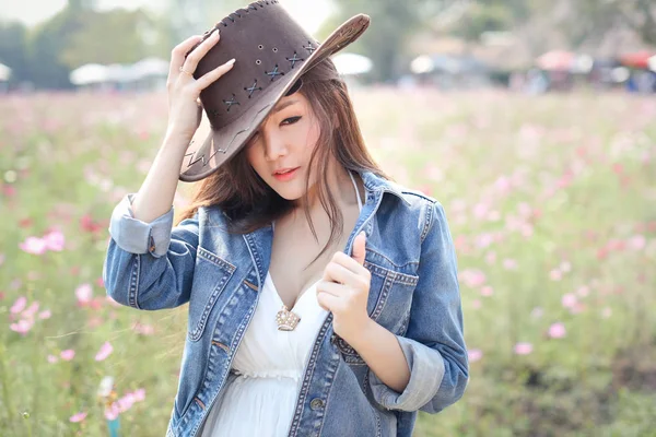 Carino e felice asiatico donna in blu jeans in piedi in natura all'aperto tra cosmo fiori campo con marrone cappello e bellezza faccia (vacanza e vacanza concetto ) — Foto Stock