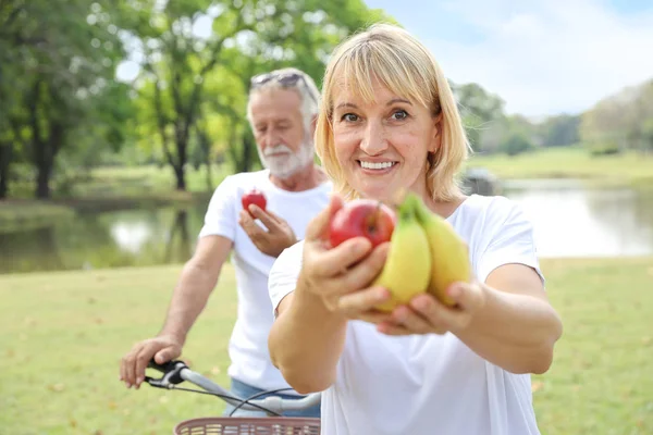 Starsza żona daje wiele rodzajów owoców do kogoś i męża trzymając jabłko na rowerze w parku w czasie letnim (zdrowa koncepcja) — Zdjęcie stockowe
