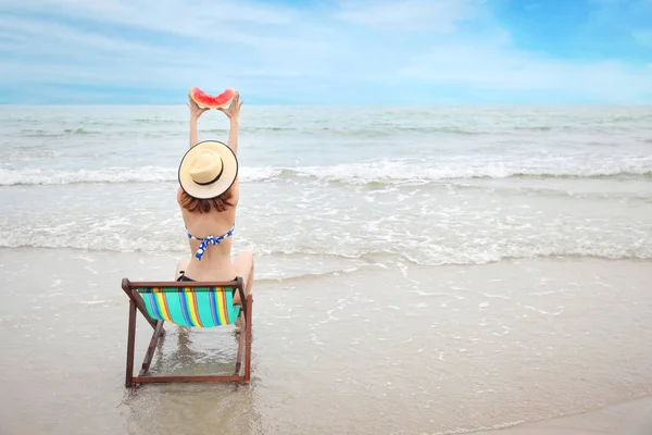 Vista trasera de mujer asiática pelo corto en un bikini azul y hermoso sombrero, ella sentada en la silla de cubierta y levantando sandía en la playa de arena con cara feliz y belleza en vacaciones de verano — Foto de Stock