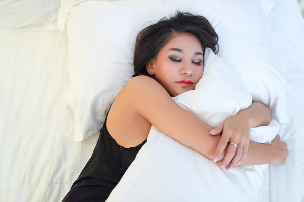 黒いかわいいアジアのモデルは、ベッドの上で眠りと抱擁白い枕を通して見る — ストック写真