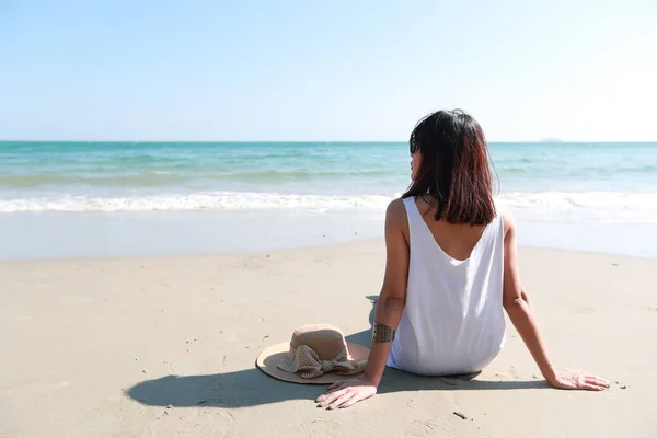 Вид сзади Портрет сексуальной азиатской молодой девушки в бикини и шляпе, глядя на тропический пляж, сидя на песчаном пляже в природе с голубым небом . — стоковое фото