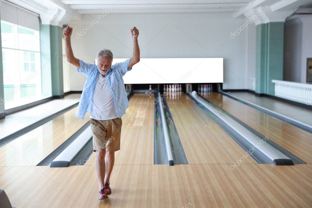 Older caucasian men, white beard and white hair in white and blue shirt playing bowling and he put his hands up after finish the game meaning of the winner in sport club