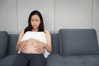 asyalı hamile kadın oturma odasında oturuyor ve midesini tutarak başka bir el vurguladı 
