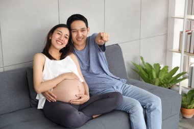 genç koca genç hamile göbek karısı kucaklayan ve oturma odasında otururken mutluluk ve gülümseme ile bir şey işaret