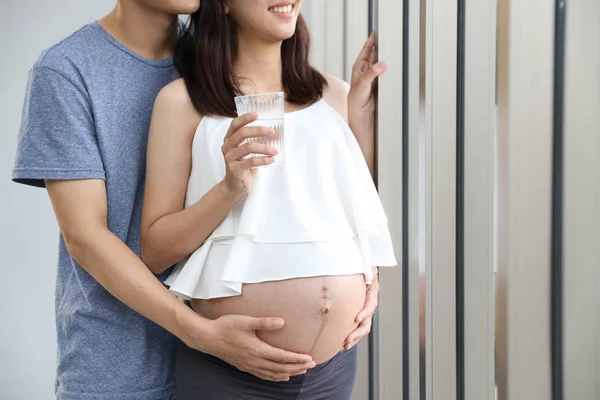 Junger Mann umarmt junge schwangere Frau mit Glück und Lächeln, während sie ein Glas Wasser in der Hand hält — Stockfoto