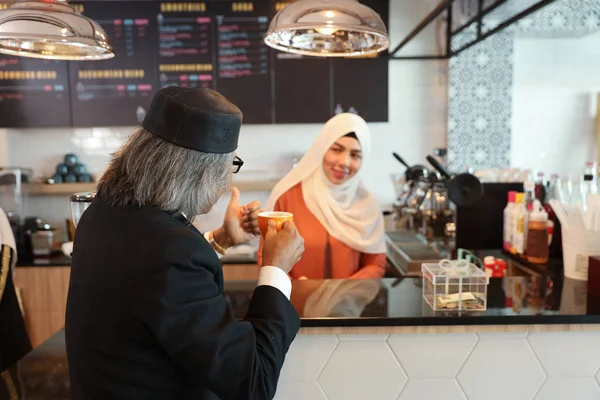 muslim customer businessman wearing black suit drinking coffee at counter and showing thumb up with young muslim barista girls background