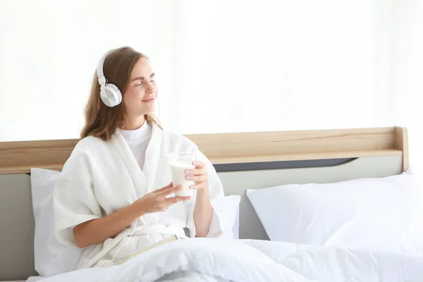 Joven mujer caucásica alegre en pijama blanco con auriculares que sostiene un vaso de leche mientras lee un libro antes de ir a dormir con la cara feliz y sonrisa en la cama en el dormitorio moderno blanco. — Foto de Stock