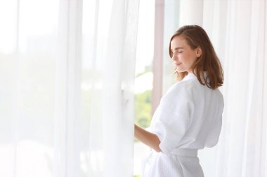 Sağlıklı ve sağlıklı bir beyaz kadın sabah uyandığında gün doğumunda uyanır ve yatak odasının penceresinin yanında mutlu bir yüzle durur.)