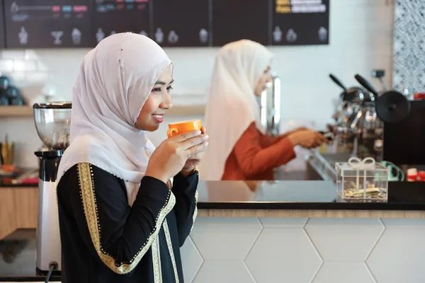 Muslim customer drinking coffee at counter with young muslim barista girl blurred background — Stock Photo, Image