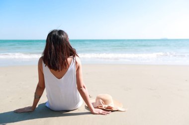 Bikini ve şapka seksi asyalı genç kız Arka görünüm portresi, mavi gökyüzü ile doğada kumlu plajda otururken tropikal plaja uzak bakıyor.
