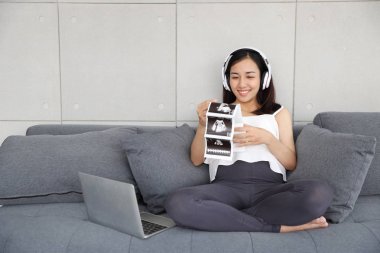 oturma odasında kanepeüzerinde ultrason fotoğrafı tutarken bilgisayar kullanarak ve müzik dinlerken güzel asyalı hamile kadın portresi 