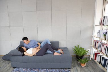 Genç Asyalı koca kanepeye uzanıp kucaklıyor ve gözleri hamile karıyla temas kuruyor aşk ve mutluluk dolu bir gülümsemeyle oturma odasında.