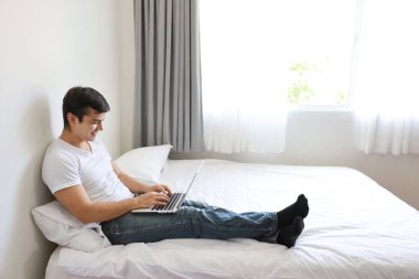 Beyaz gömlekli ve mavi kotlu yakışıklı beyaz adamın yatak odasındaki bilgisayarı rahatlamış ve gülümseyen yüzüyle kullanmasının portresi.