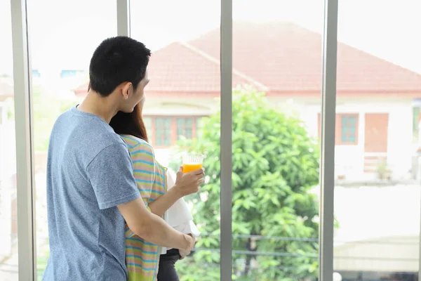 Back view image of young asian husband embracing young asian pregnant belly wife and looking at something through the window with happiness and smile while her holding orange juice — Stock Photo, Image