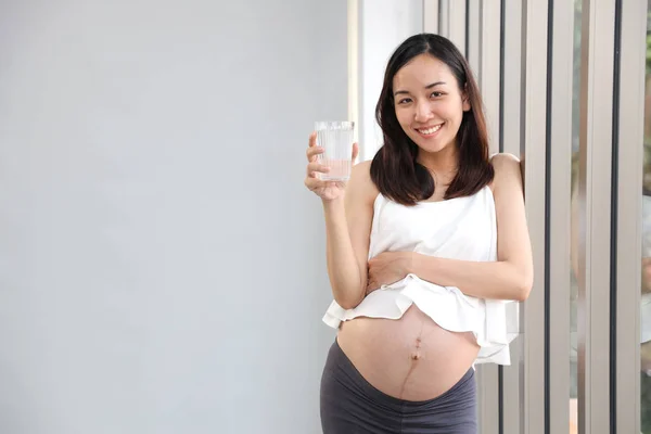 Joven asiático saludable embarazada vientre sonrisa mientras sostiene un vaso de agua con feliz cara y ojos contacto izquierda copia espacio (salud cuidado concepto ) —  Fotos de Stock
