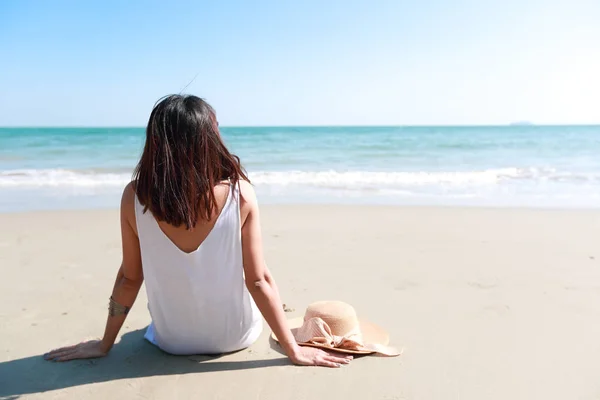 Вид сзади Портрет сексуальной азиатской молодой девушки в бикини и шляпе, глядя на тропический пляж, сидя на песчаном пляже в природе с голубым небом . — стоковое фото