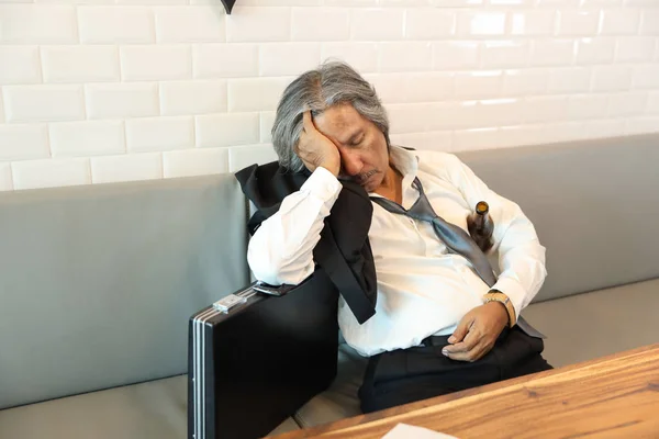 Senior-Geschäftsmann mit Anzug, der eine leere Alkoholflasche in der Hand hält und auf dem Sofa im Café schläft (Stress oder Misserfolg im Geschäftskonzept)) — Stockfoto