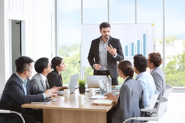 caucasian lecturer giving public presentation with company business graph result on white board in meeting room and multiethnic business people are paying attention (training or seminar concept)